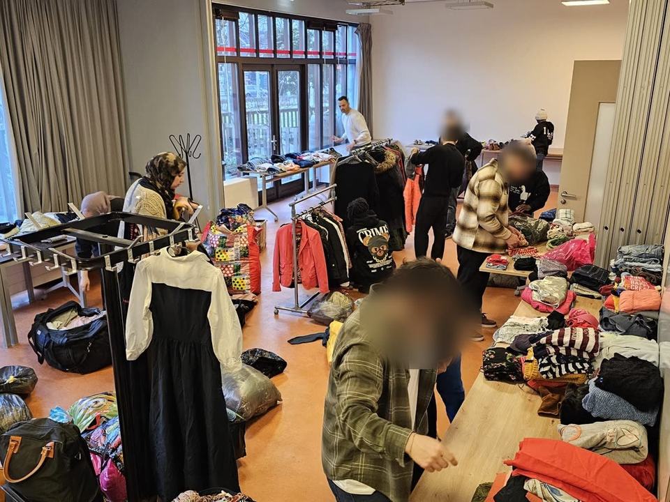 Yara Gamal, onze oprichter, en onze medeoprichter sorteren met vrijwilligers gedoneerde kleding op tafels en kledingrekken in een buurtzaal, met zakken vol donaties op de vloer. Sommige gezichten zijn vervaagd.