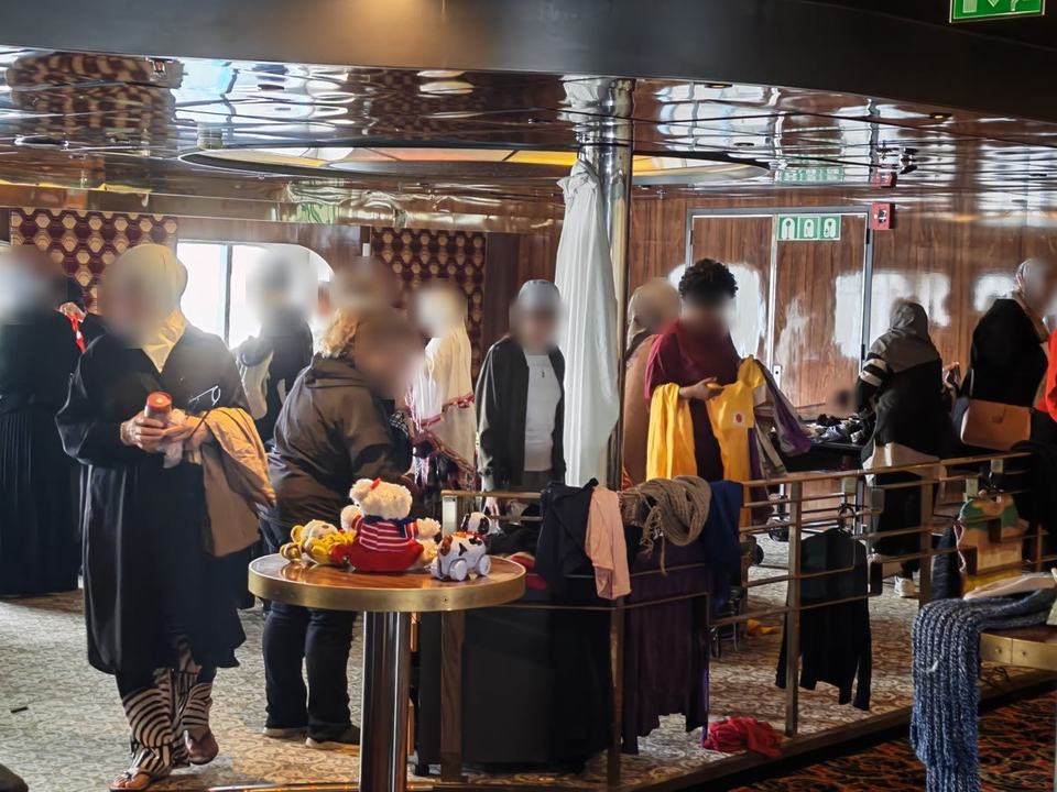 Vrouwen kijken naar kleding en speelgoed op ronde tafels en rekken in een lounge aan boord van een schip. Gezichten zijn vervaagd.