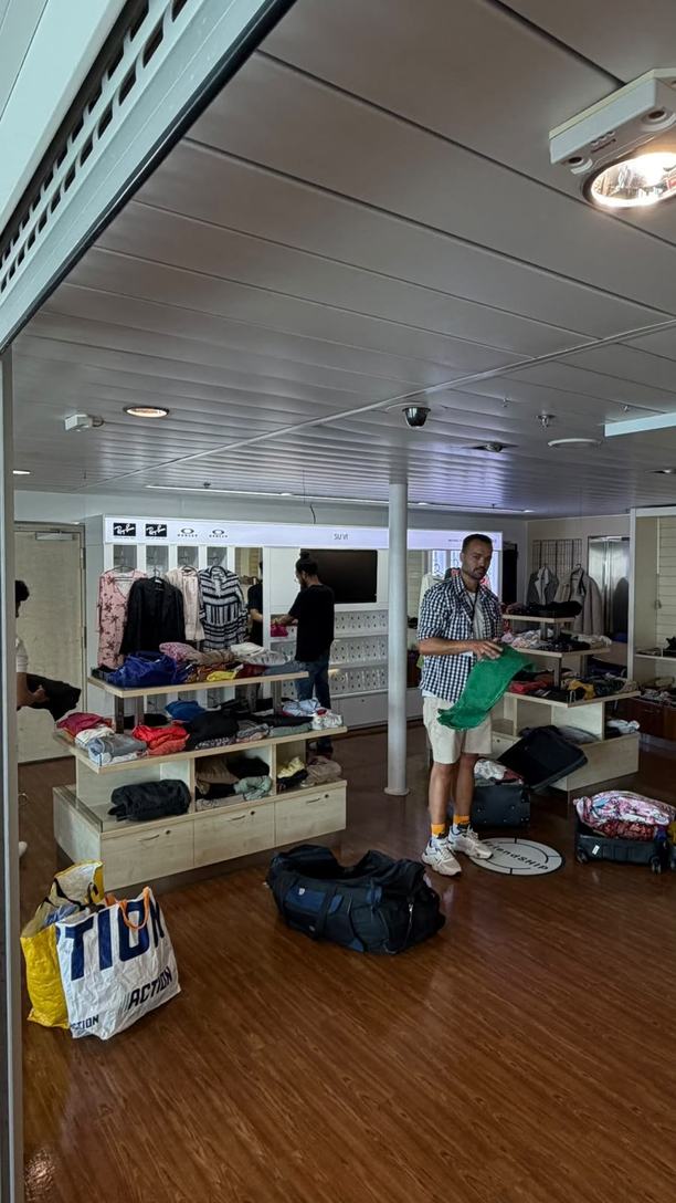Een winkelruimte op een schip met schappen en rekken vol kleding, tassen op de houten vloer, en onze medeoprichter en een vrijwilliger die kleding klaarleggen.