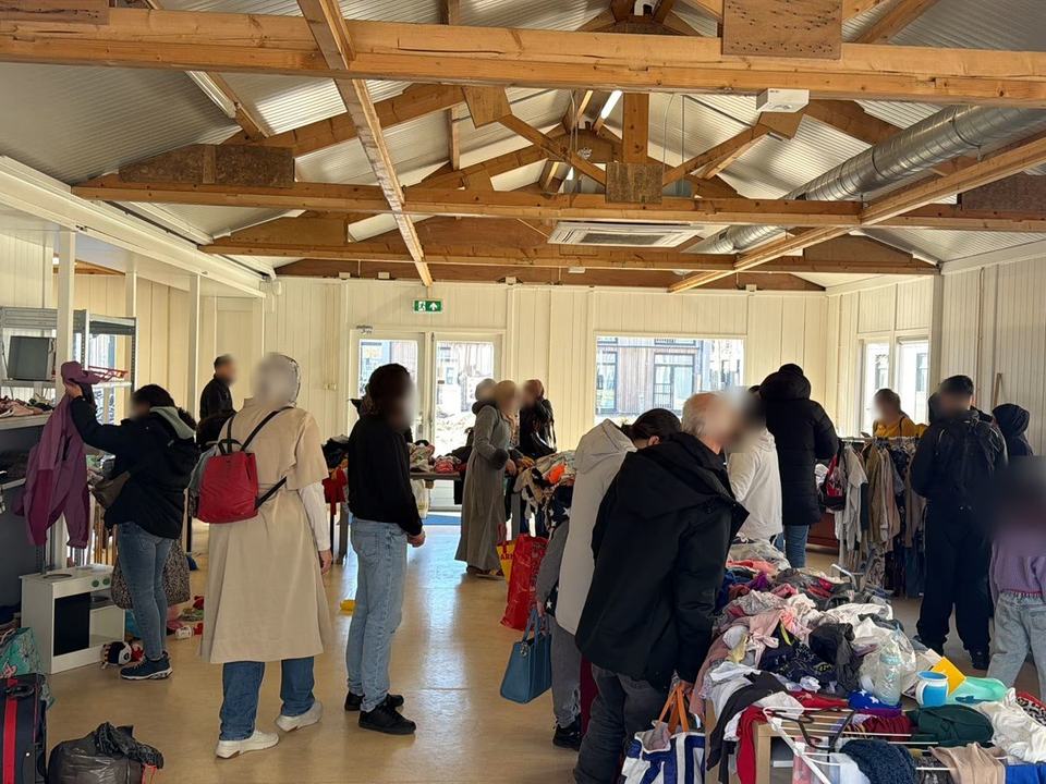 Mensen kijken rond bij tafels en schappen met kleding, schoenen en speelgoed in een grote zaal met een houten dak.