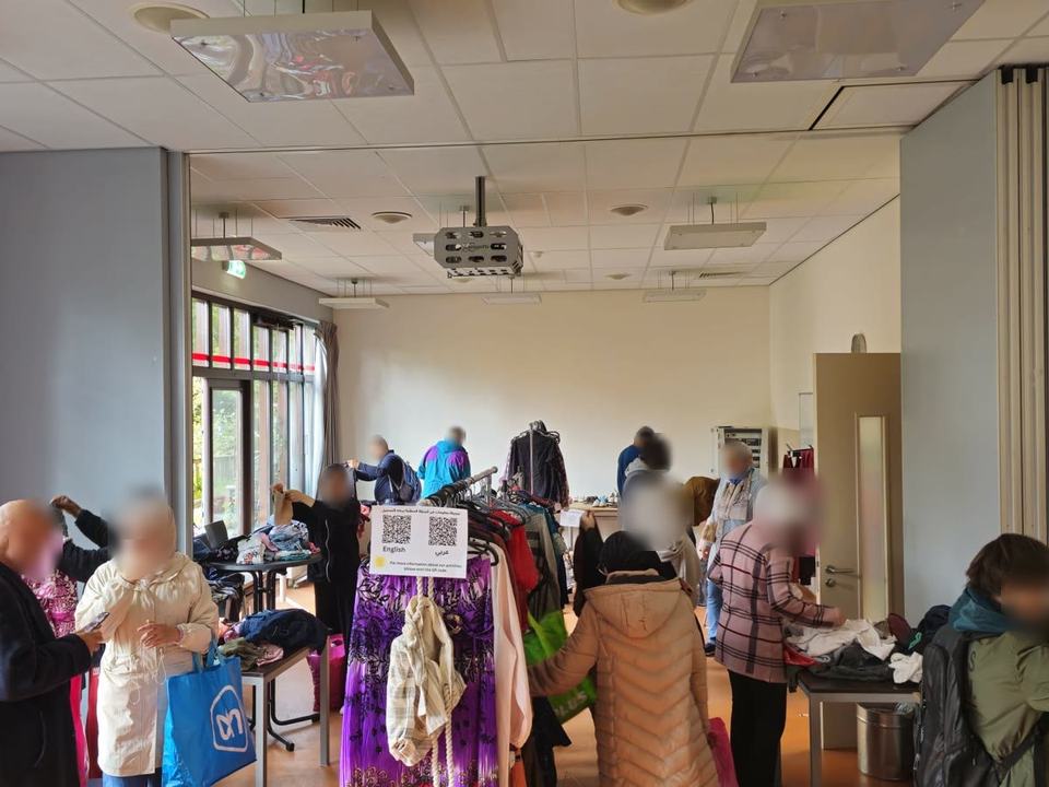 Mensen kijken rond bij rekken en tafels met gedoneerde kleding in een grote zaal.